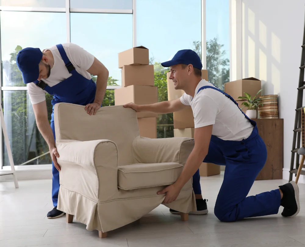 Two professional movers in blue overalls adjusting a beige armchair in a bright room with moving boxes.