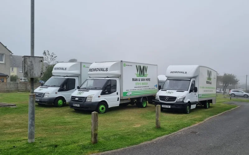 Fleet of VM Removals vans parked on a grassy area, showcasing white vehicles with green accents and branding for moving services.